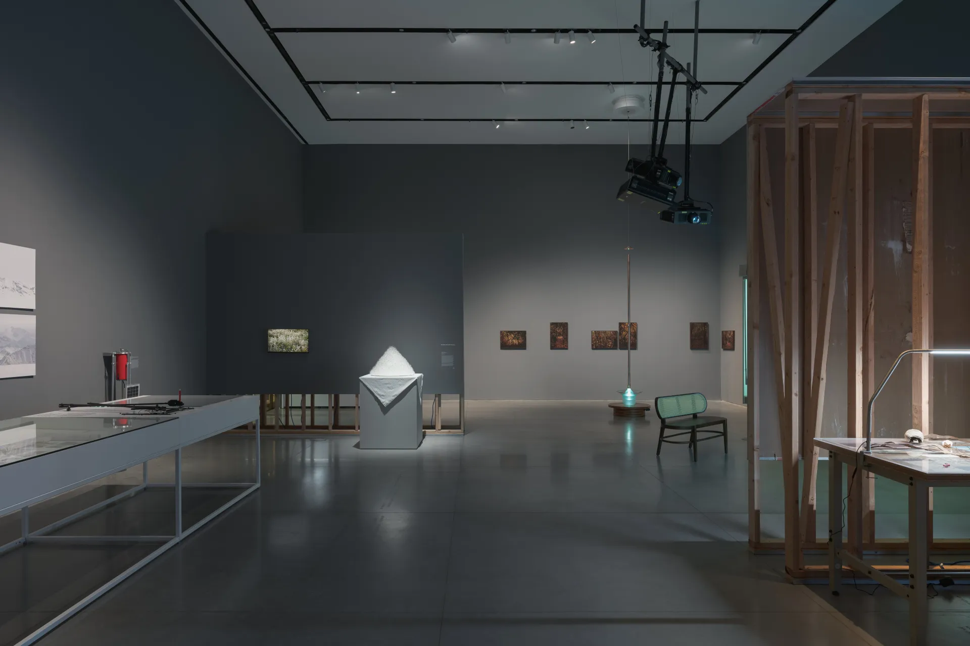 A gallery exhibition showcasing several artworks such as a metal table, a small monitor, a soft white sculpture, framed prints, a glass bowl, projection equipment hanging from the ceiling, and a rattan bench.