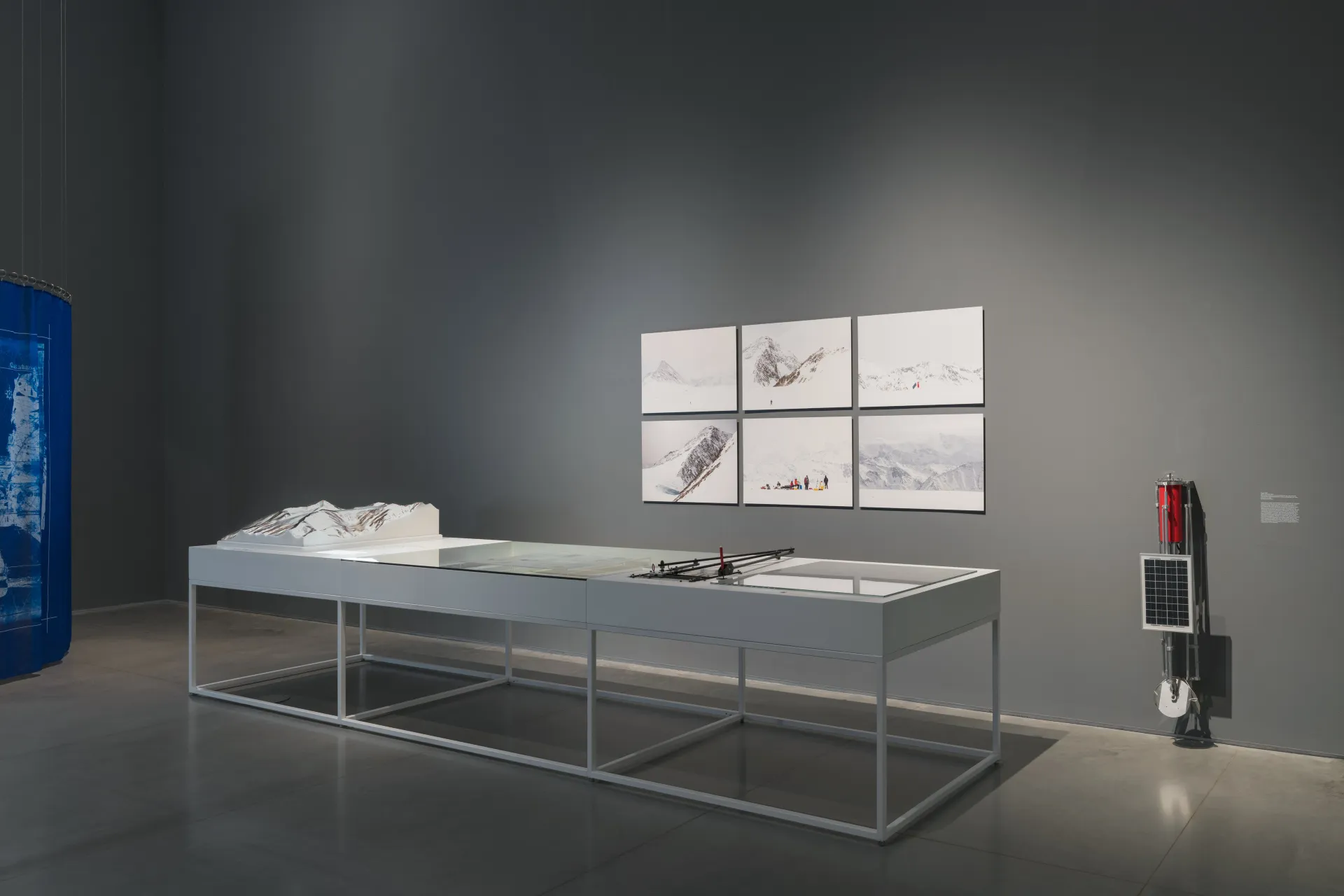 A diagonal view of a long metal table with a three-dimensional plaster model of a glacier. Six images of snowy alpines are hung behind the table. And a satellite sculpture stands to the right of the table