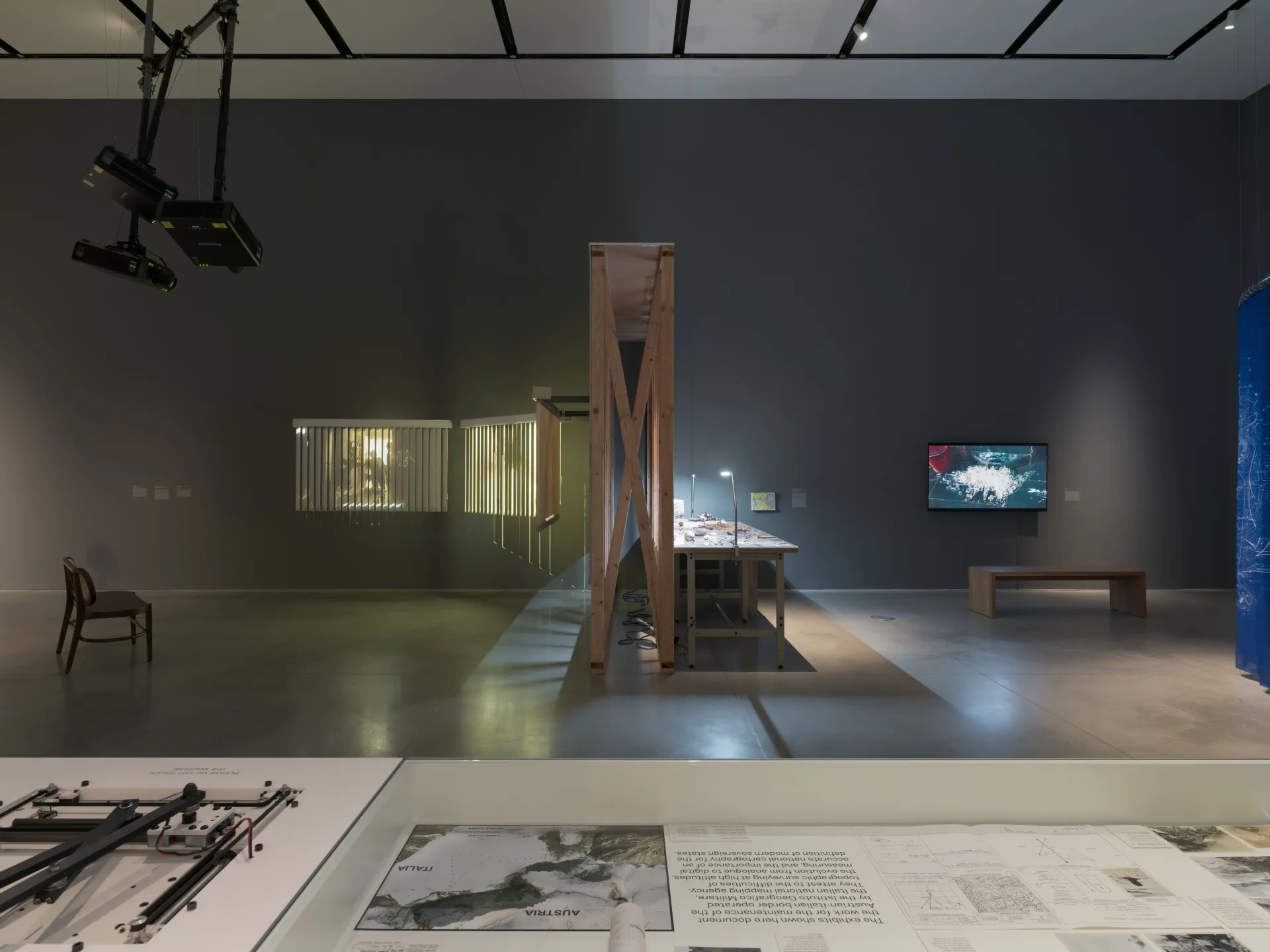 A snapshot of a gallery exhibition with the perspective above a metal and glass research table. Benches, projections, a side of a wooden wall, a lit-up table with found objects, and a video monitor are displayed in the distance.