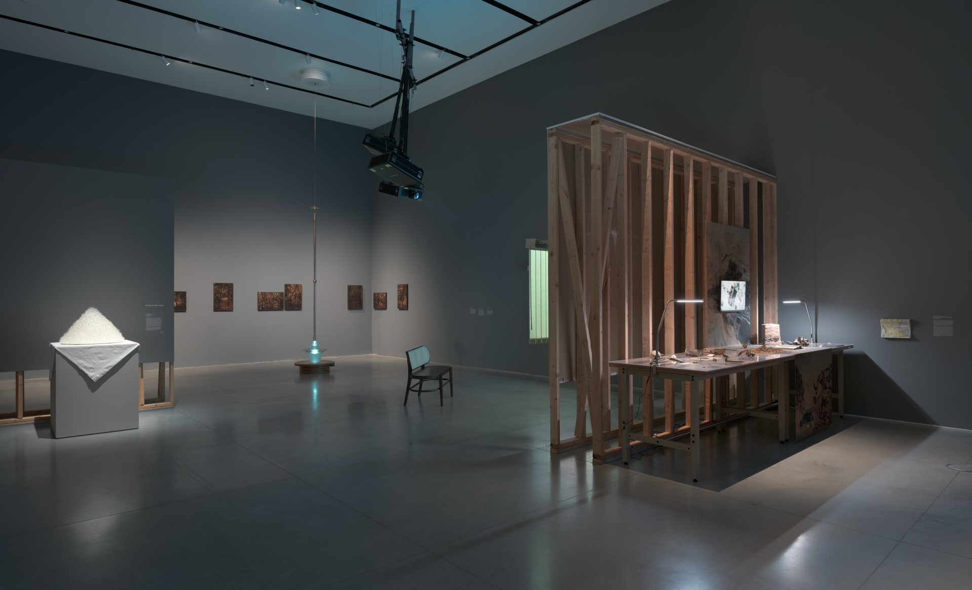 An exhibition with several artworks such as a white pyramid soft-sculpture, photographs, a glass bowl, and an industrial table with found objects.