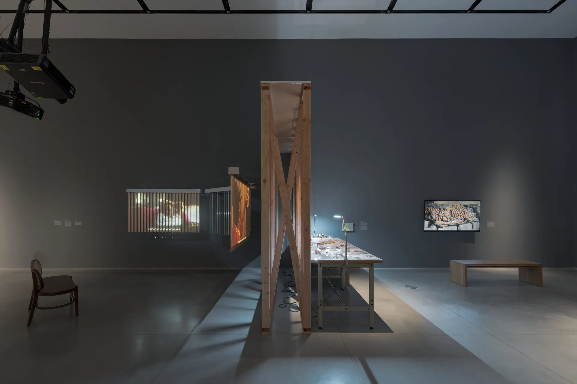 A gallery view of several benches, projections, a side of a wooden wall, a lit-up table with found objects, and a video monitor.