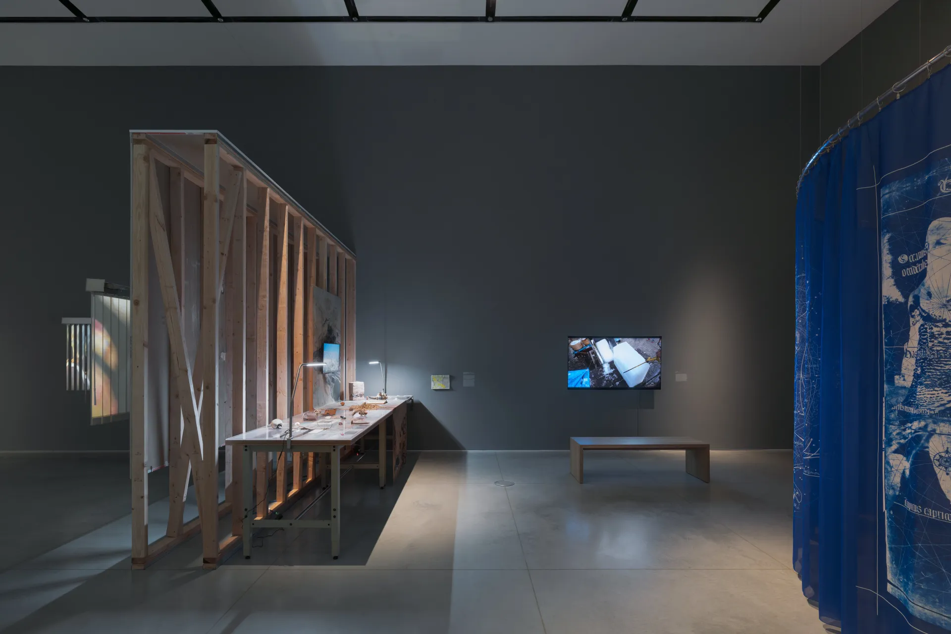 An exhibition displaying an industrial table with found objects against an unfinished wall construction, a video monitor and wood bench, and corner of a curved blue tapestry.