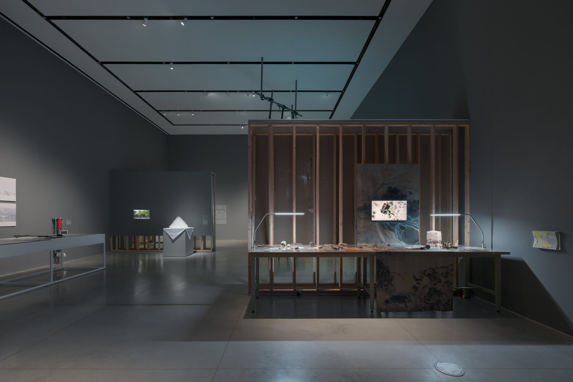 A gallery exhibition featuring multiple industrial tables with archival and research objects. One table is against an unfinished wall construction. A soft-sculpture of a white pyramid is seen in the back.