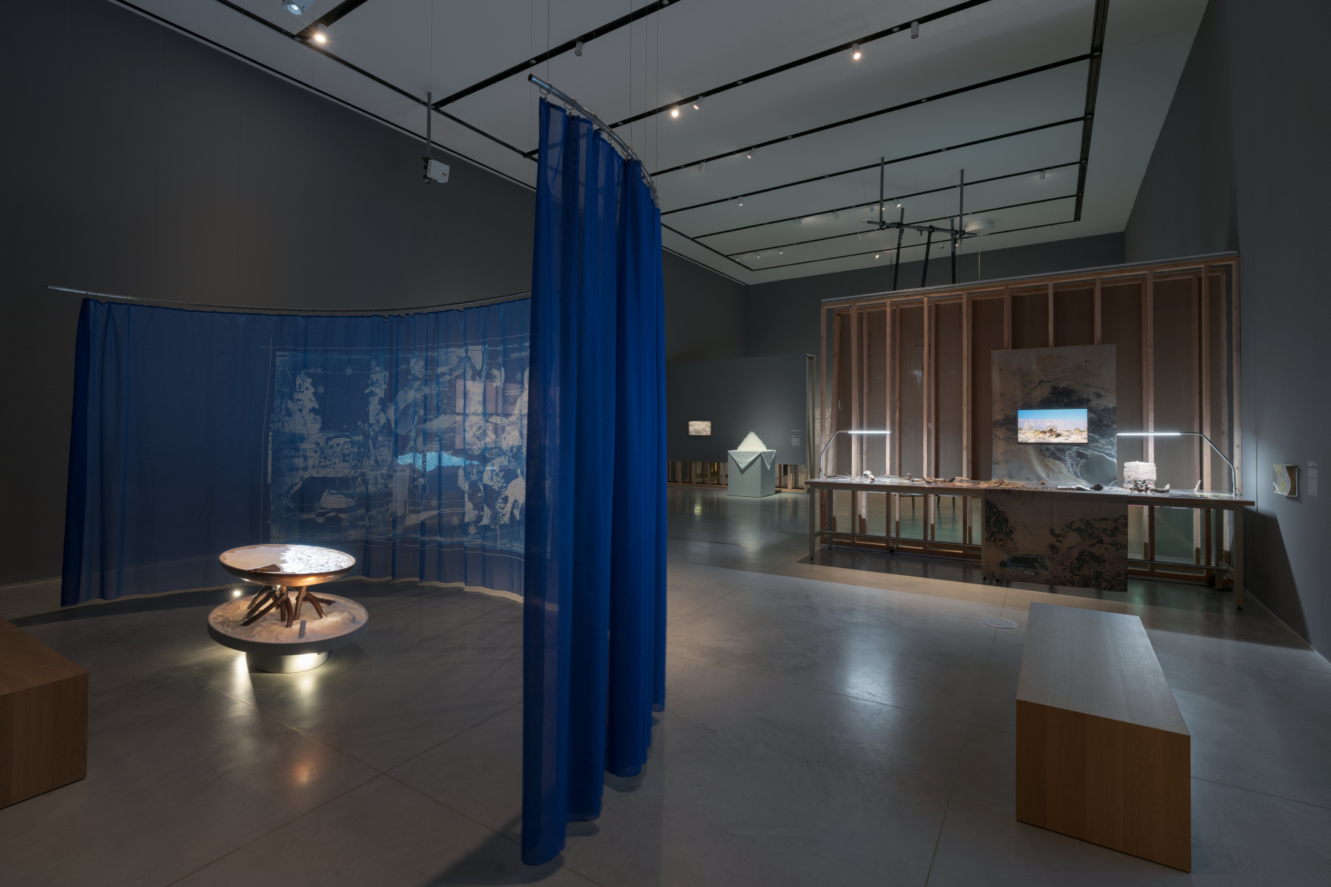 A diagonal view of an art show displaying a projection onto an illuminated table full of sand, surrounded by a translucent blue tapestry. There are several benches, and multiple artwork installations in the distance.