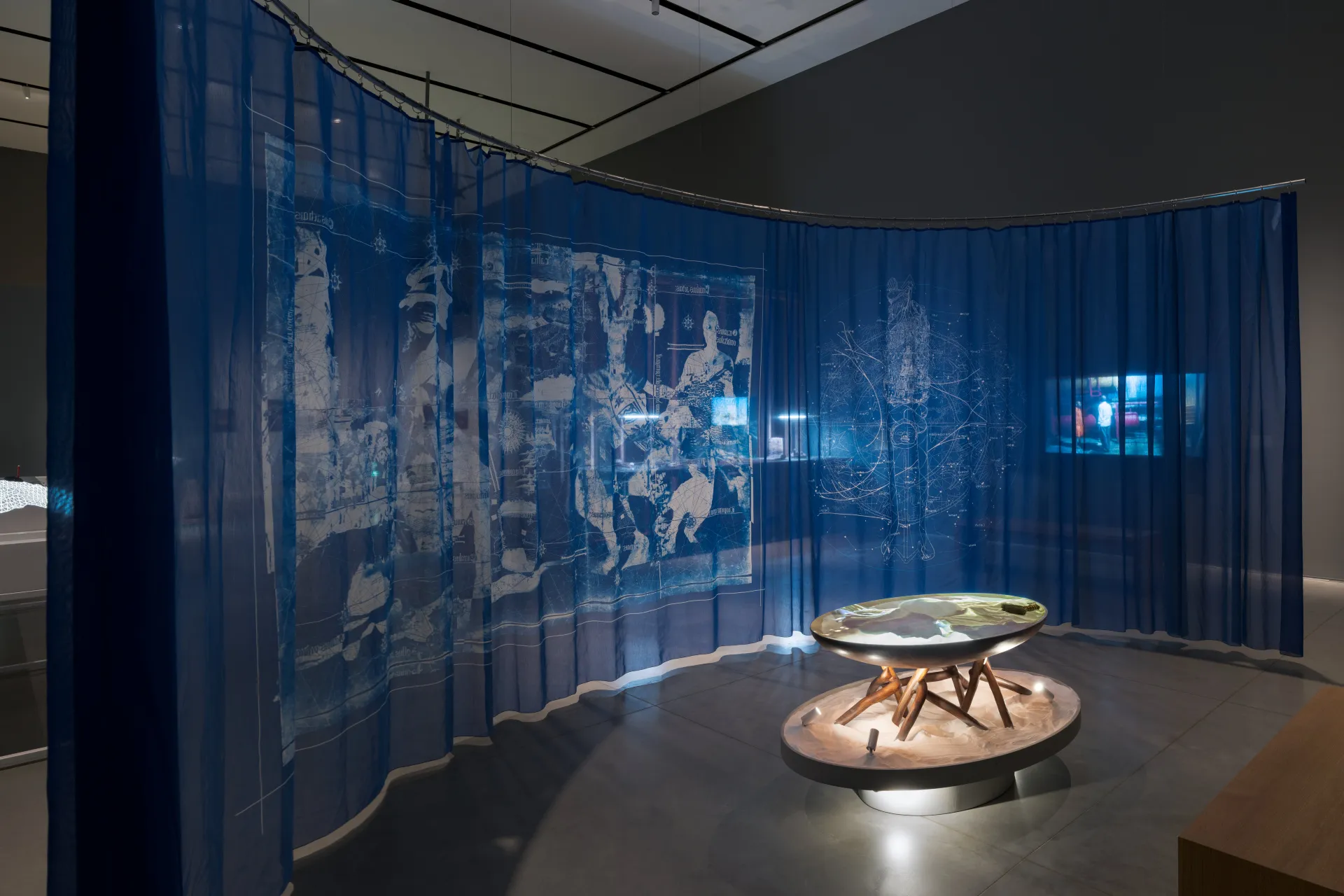 A translucent blue tapestry curved in a moon like shape, creating an intimate room. A wooden table on top of an oblong pedestal stands in the center. The table, full of sand, shows a video projection.