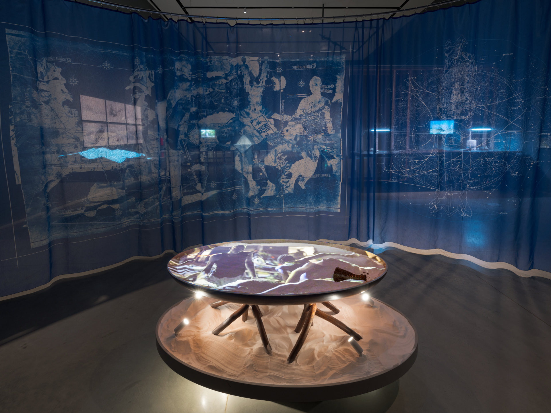 A sheer blue tapestry serves as the backdrop for an organically-shaped wooden table with branch-like legs, resting in sand. A video is projected onto the sand on the surface of the table.