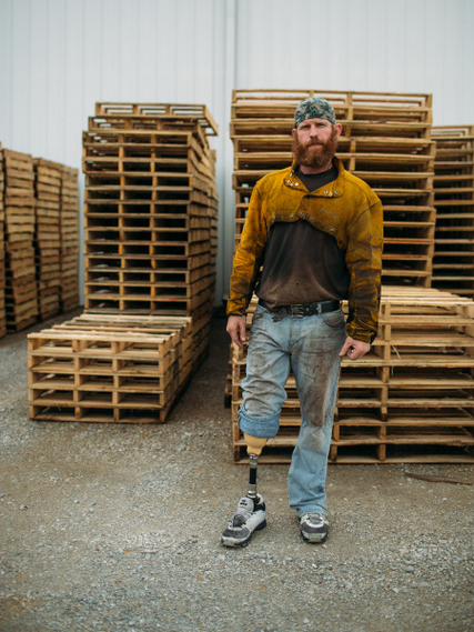 Billy Brimblecom, Jr, man with a red beard and a prosthetic leg stands in front of stacks of wooden pallets. He is wearing a yellow work jacket and a camouflage bandana.