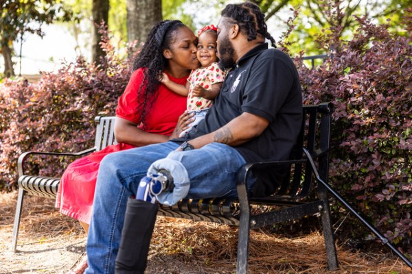 A Black man with a prosthetic leg sits on a park bench with his partner and young daughter. The family shares a loving moment outdoors.