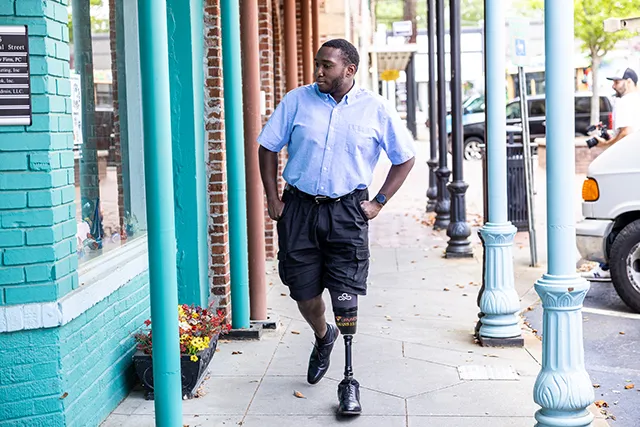 A Black man with a prosthetic leg walks down a city sidewalk lined with teal columns. He is wearing a blue button-down shirt and black shorts.