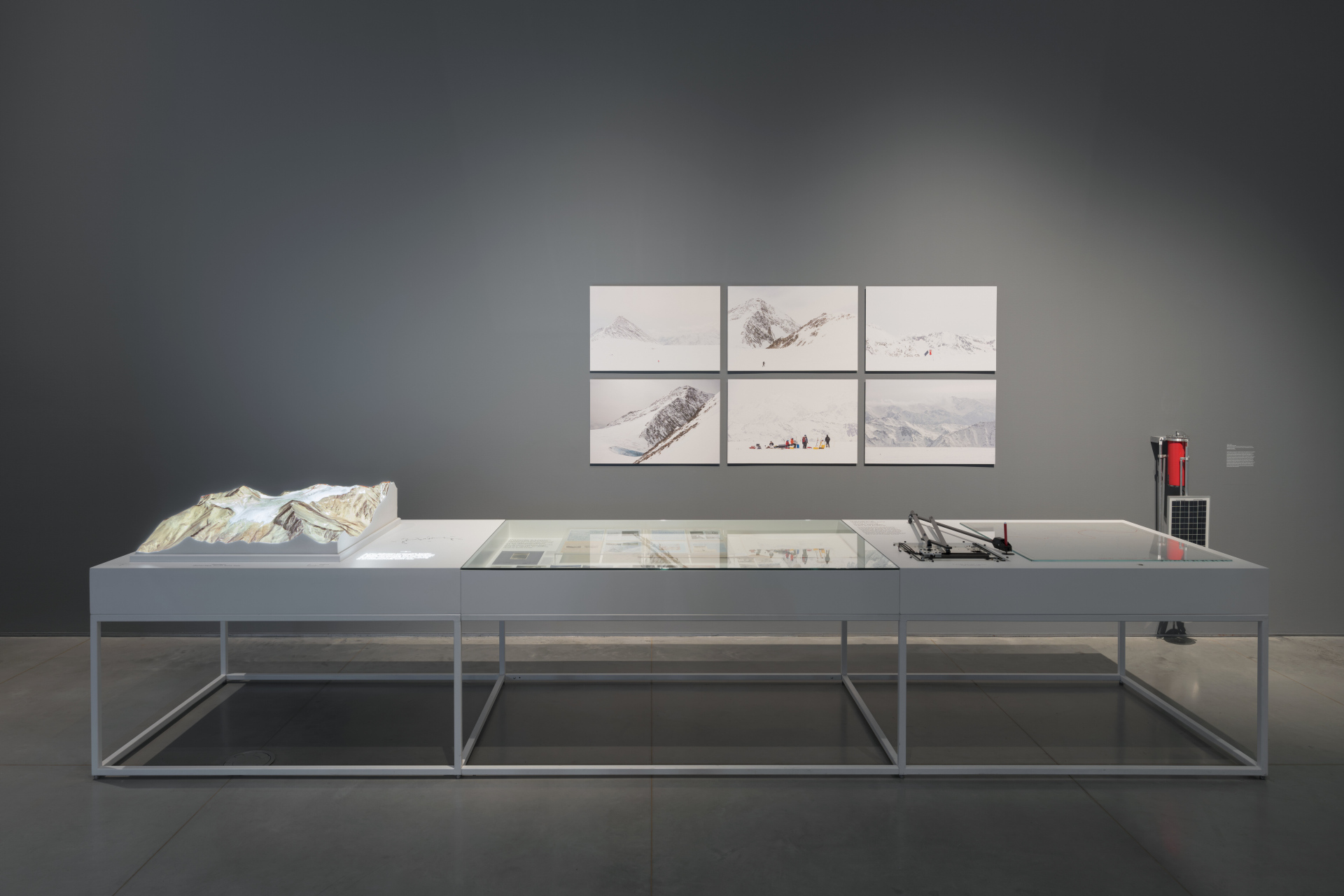 A long metal table with various research media, a three-dimensional plaster model of a glacier, and photographs of snowy mountain ranges.