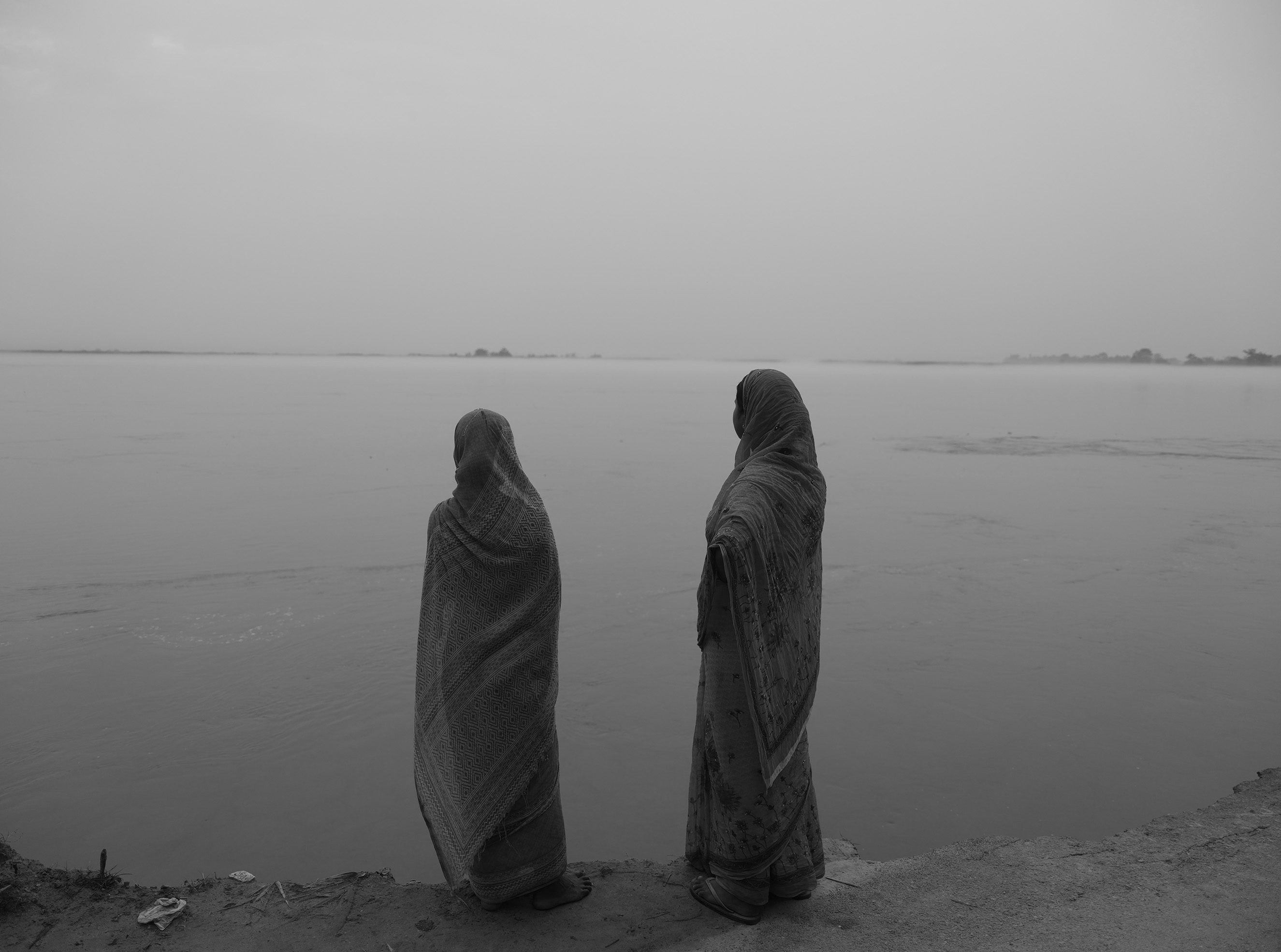 Two women wearing long headscarves and dresses stand together, looking out over a large body of water.