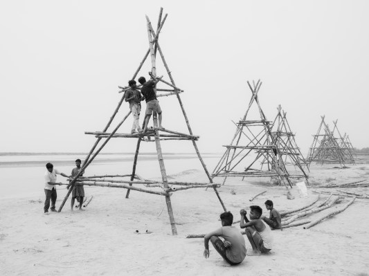 A gray-scale photo of South Asian teenage boys building pyramid structures out of bamboo sticks on a beach. Some boys are sitting down taking a break, and some are climbing and working on the structures.
