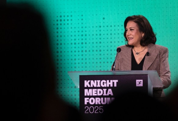 Maribel Pérez Wadsworth, a woman with brown hair in a checkered blazer speaks at a podium during the Knight Media Forum 2025. She is smiling, standing against a vibrant green textured backdrop.