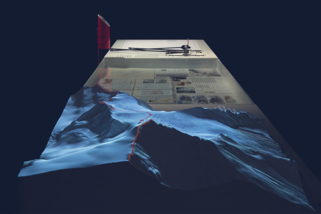 A 3-D topographical model showcasing a mountainous terrain with a red path, illuminated against a dark background, and a desk with several documents and tools.