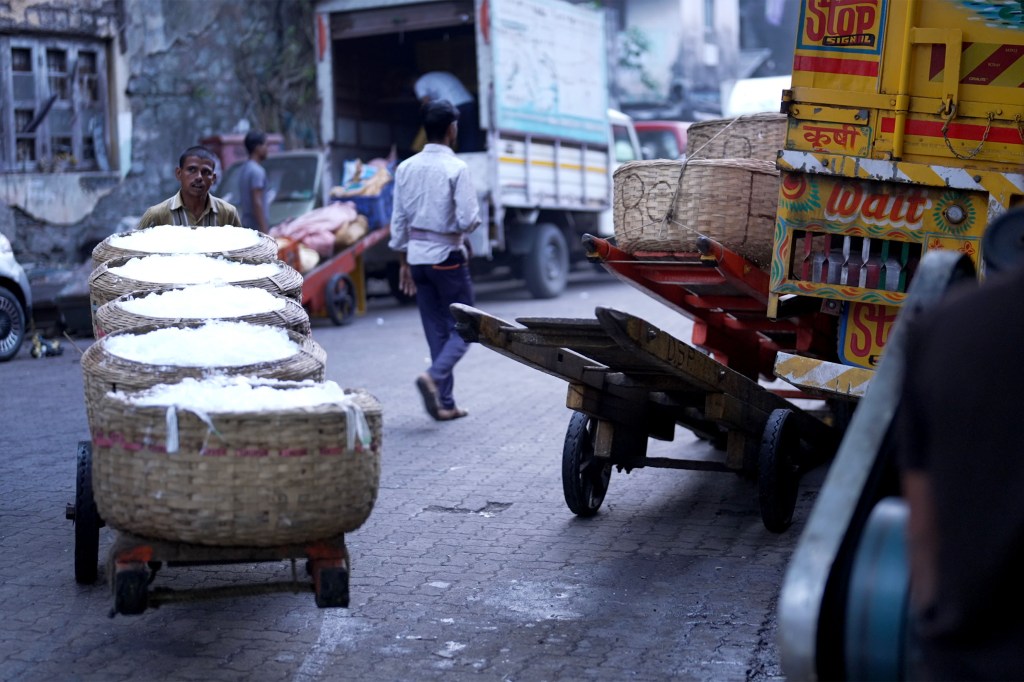A bustling street scene with a man pushing a cart of large baskets filled with ice. Vehicles and more baskets are seen in the busy and industrious background.