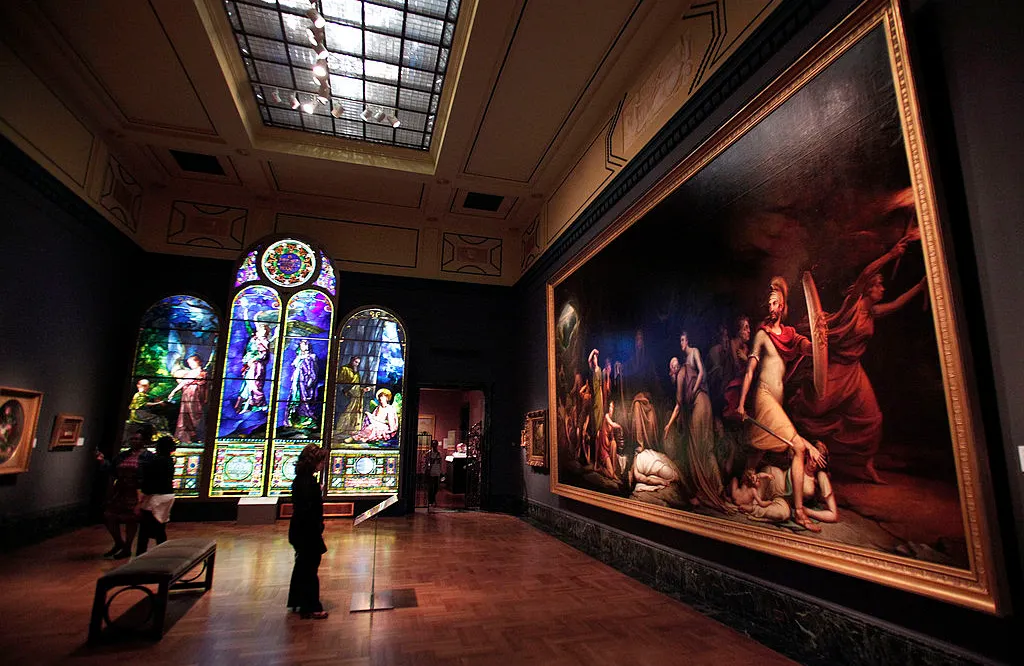Visitors in a dimly lit gallery at the Detroit Institute of Arts view large-scale artworks, including vibrant stained glass windows and a massive classical painting. This image showcases museum tourism, art history education, and cultural heritage preservation in a world-class institution.
