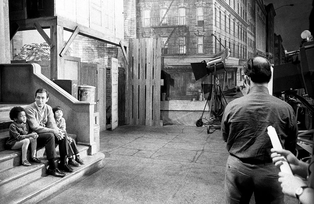 A black-and-white historical photograph shows a man and two children sitting on the front steps of a brownstone building set. A camera operator and film equipment are visible in the foreground, capturing a behind-the-scenes moment of television production and mid-century urban storytelling.