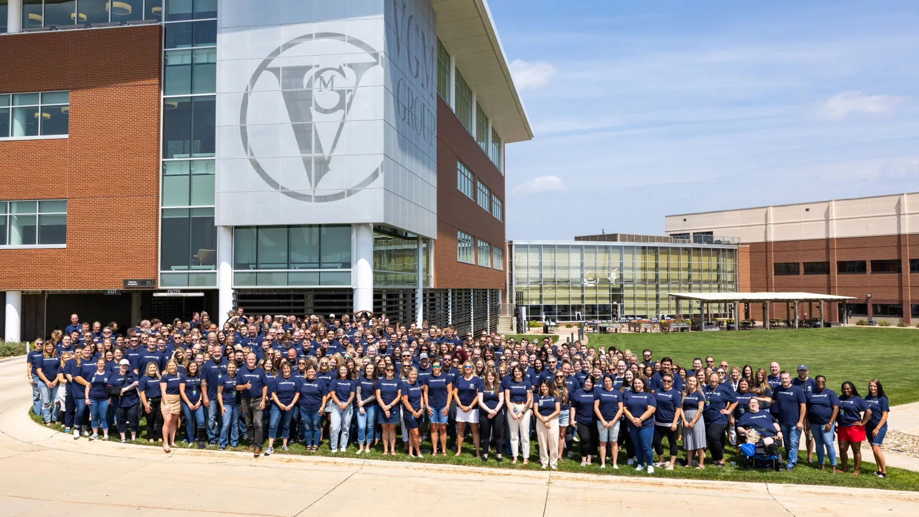 A large group of hundreds of employees in matching navy blue t-shirts pose for a panoramic outdoor photo in front of the modern, brick and glass VGM Group headquarters. This image represents corporate culture, employee engagement, and large-scale team building in a professional setting.