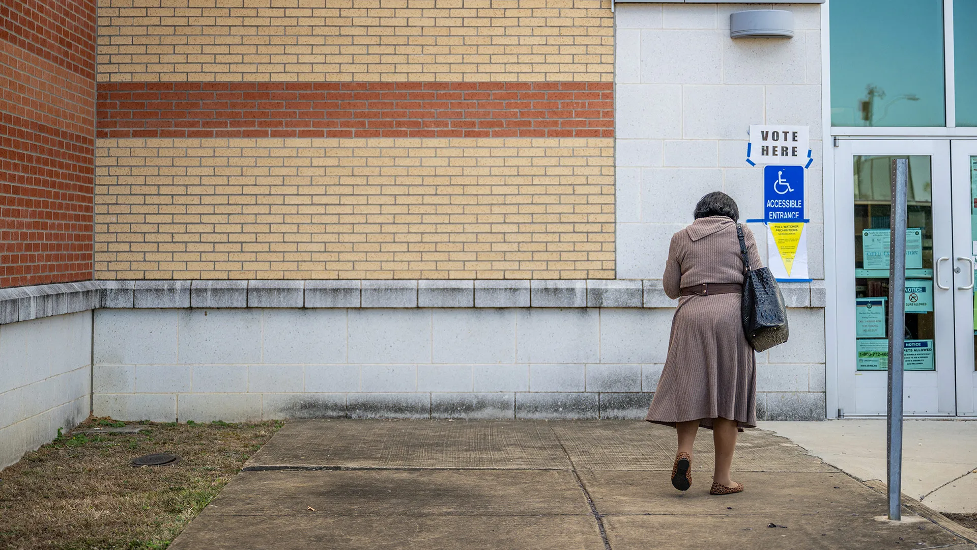 A woman from behind, carrying a black tote bag and wearing a brown dress, walks toward a building entrance. A "VOTE HERE" sign and an accessibility symbol are posted next to the door. This image represents voter participation, civic engagement, and accessible polling locations.