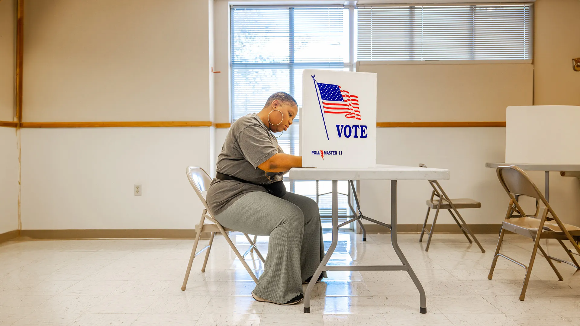A woman sits at a white folding table inside a polling station, focused on filling out her ballot behind a "VOTE" privacy screen featuring the American flag. The setting is a clean, brightly lit room with large windows and additional voting booths in the background.