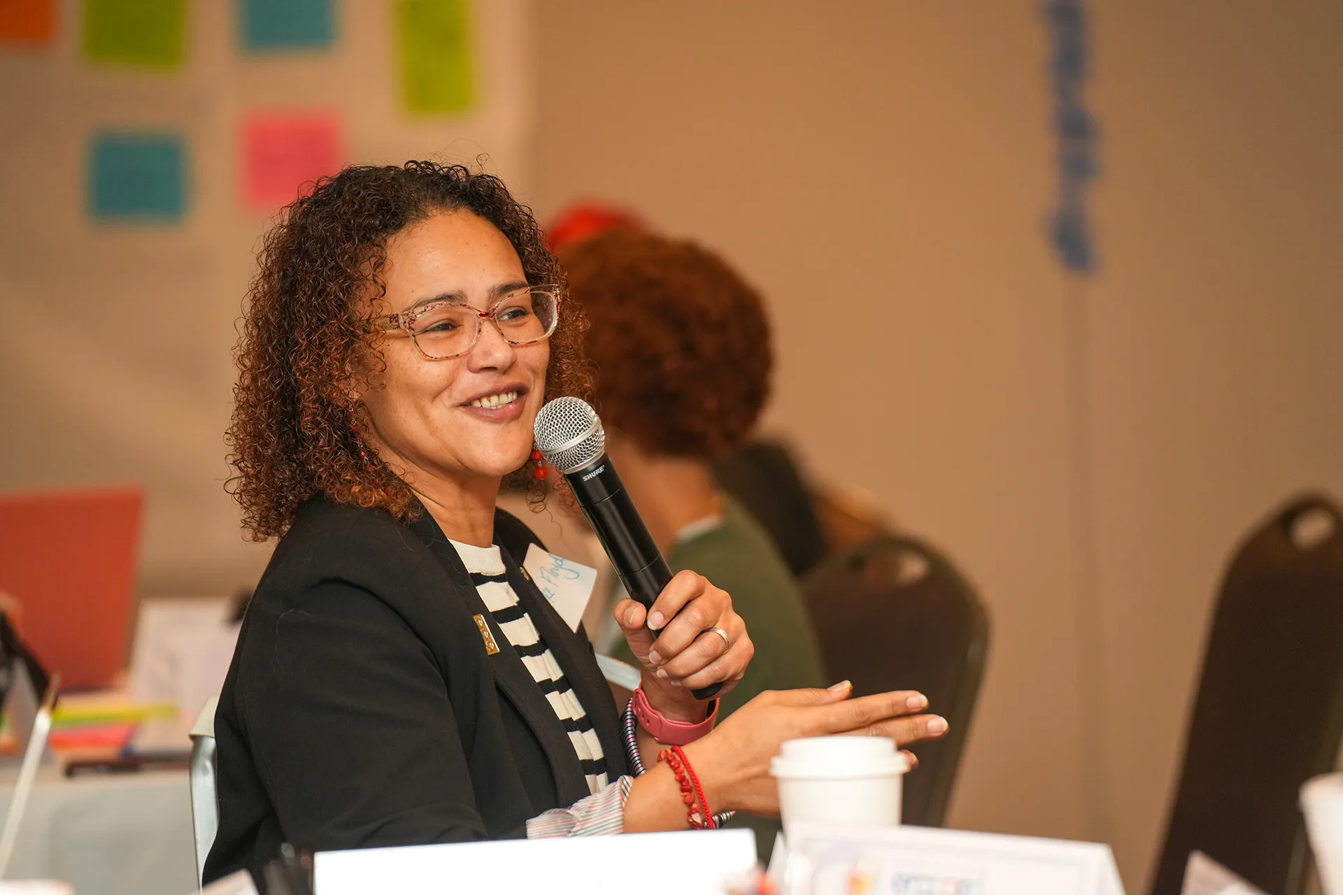 A smiling woman with curly hair and glasses holds a microphone while speaking at a community workshop. In the background, a wall is decorated with colorful sticky notes, symbolizing active civic engagement, collaborative brainstorming, and grassroots leadership in a professional meeting setting.