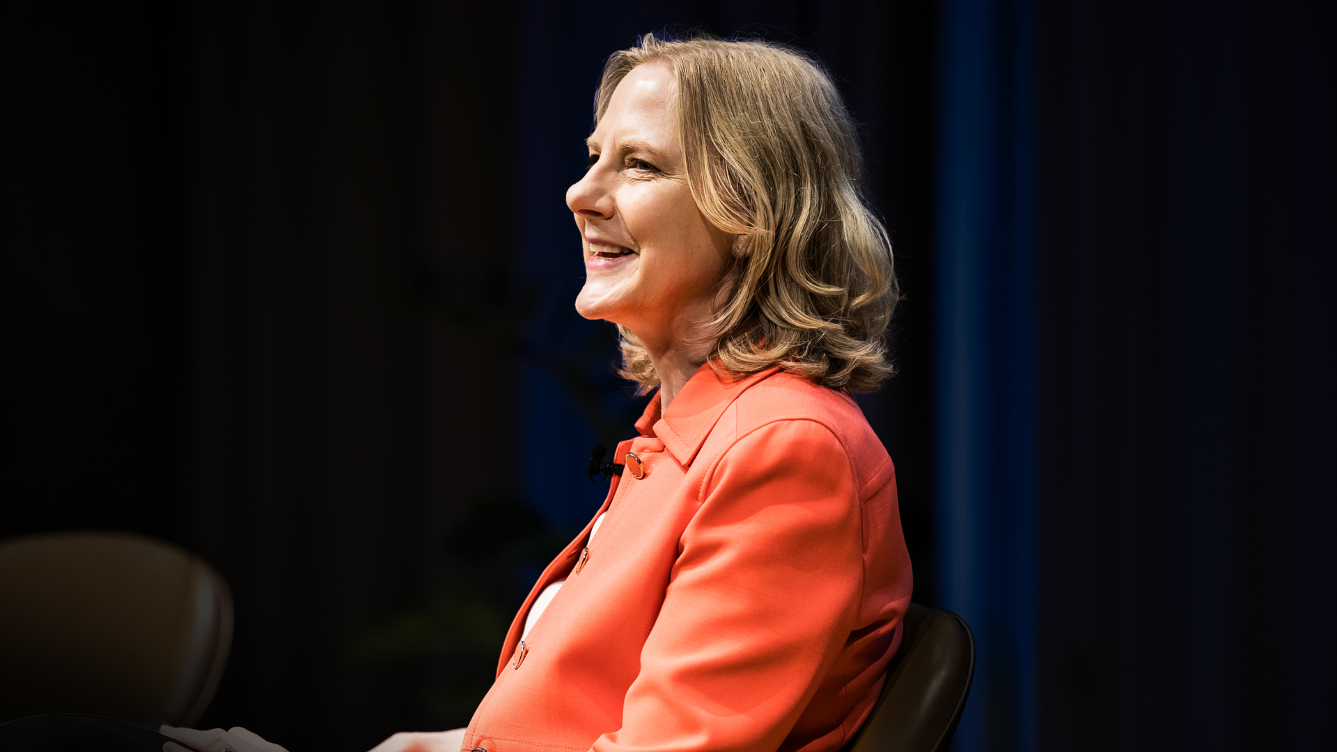 Profile portrait of a smiling woman, Heather Gerken, with blonde hair, wearing a bright coral blazer, sitting on a stage during a speaking engagement. The background is dark with a subtle blue curtain.