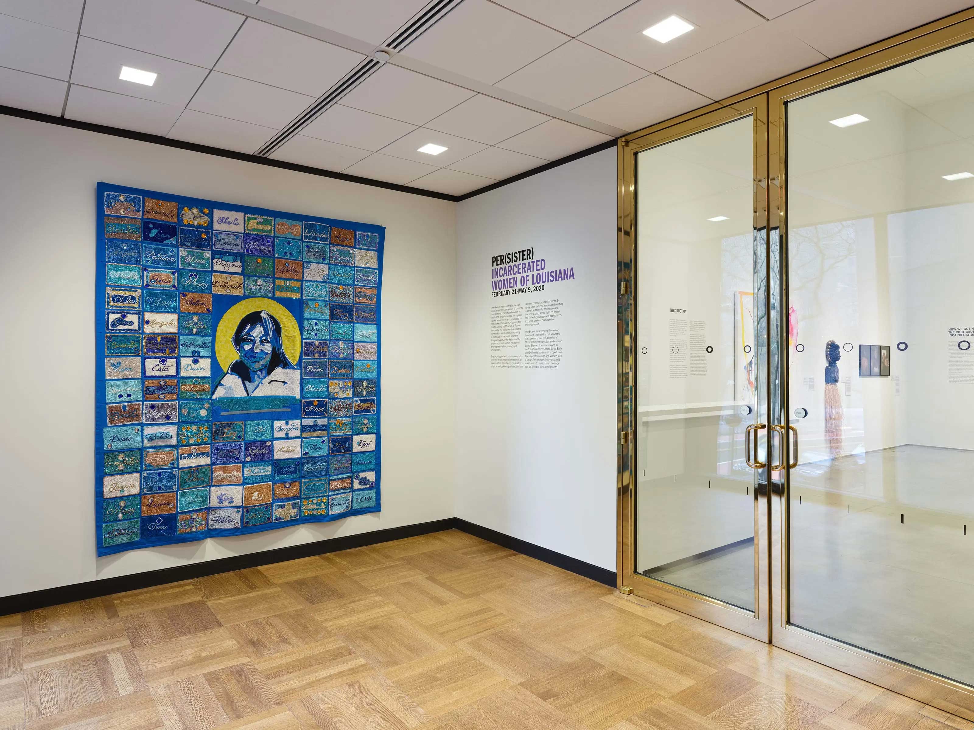 Entrance to the Ford Foundation Gallery exhibition, "Per(sister): Incarcerated Women of Louisiana." A large blue quilt featuring a yellow-haloed portrait hangs on a white wall next to gold-trimmed glass doors.