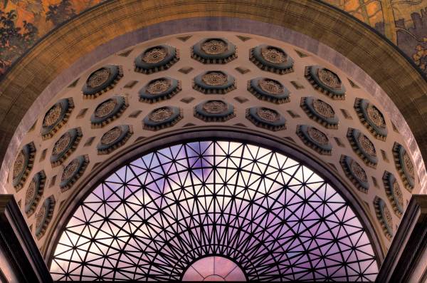 A low-angle shot of a grand architectural dome featuring an intricate, coffered ceiling with teal and gold floral patterns. A large semi-circular arched window with a geometric metal grid looks out to a soft sky.