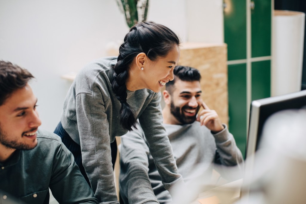 Three young professionals in casual attire work collaboratively in a modern office. A woman with a long braid smiles while looking at a computer screen alongside two male colleagues.