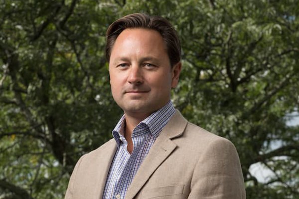 Headshot of Jason W. Ingle, wearing a tan blazer over a blue and white checkered shirt. He is posing outdoors against a lush, soft-focus background of green trees.
