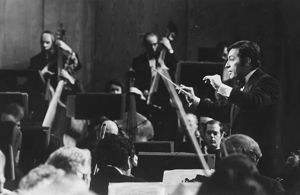 A historical black-and-white photograph of a conductor leading an orchestra. The conductor is in mid-motion, holding a baton, while musicians with double basses and other instruments are visible in the background.