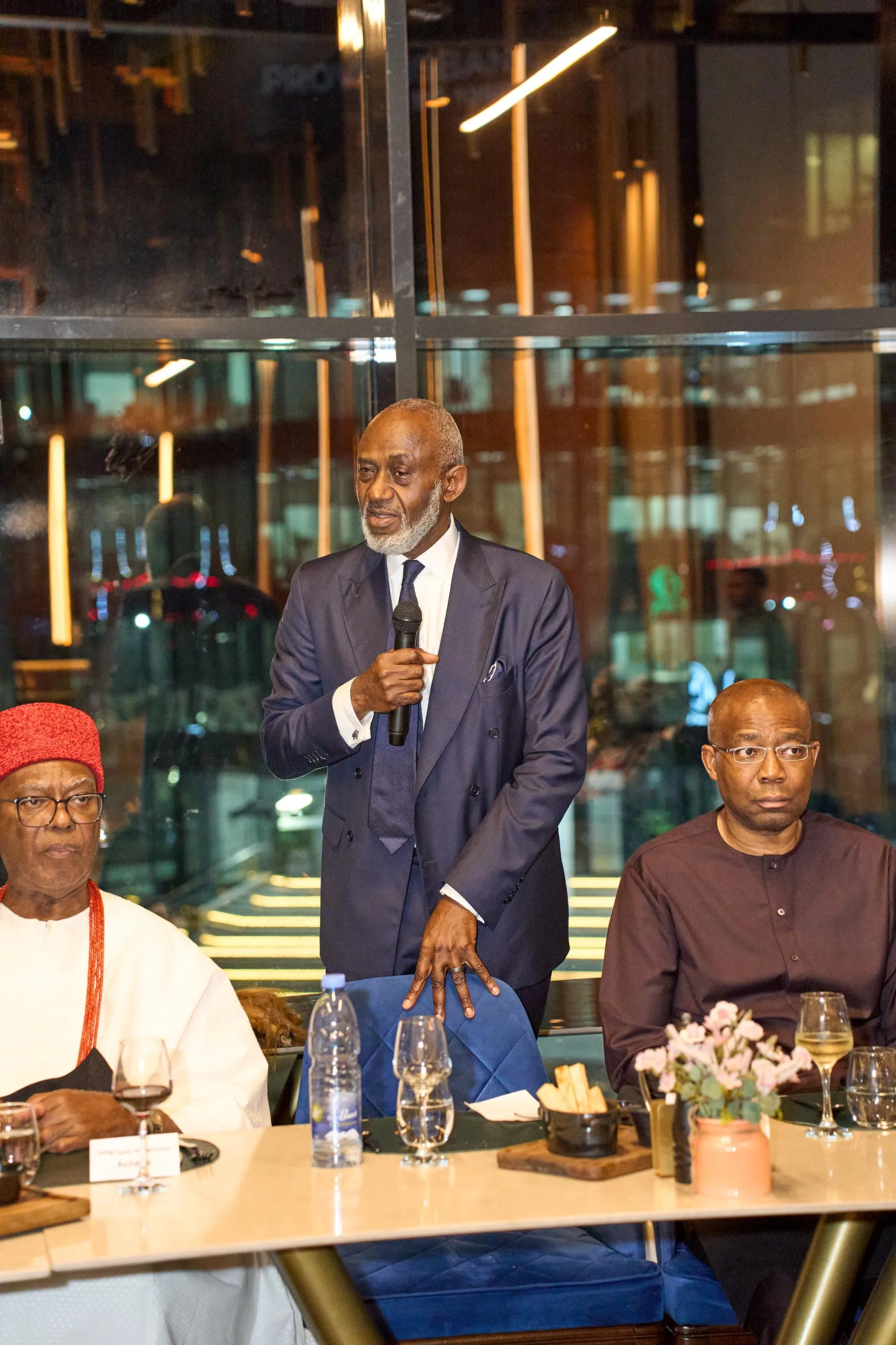 A distinguished man in a navy blue suit speaks into a microphone at a formal dinner event in Nigeria. Flanked by other seated leaders, including one in a traditional red cap, this image illustrates strategic dialogue, high-level networking, and leadership collaboration.