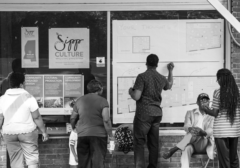 A black-and-white photo shows community members at a Sipp Culture storefront in Mississippi. A man writes on a large architectural plan titled "Your Thoughts?" while others observe, highlighting community-engaged design, rural development, and local civic participation.