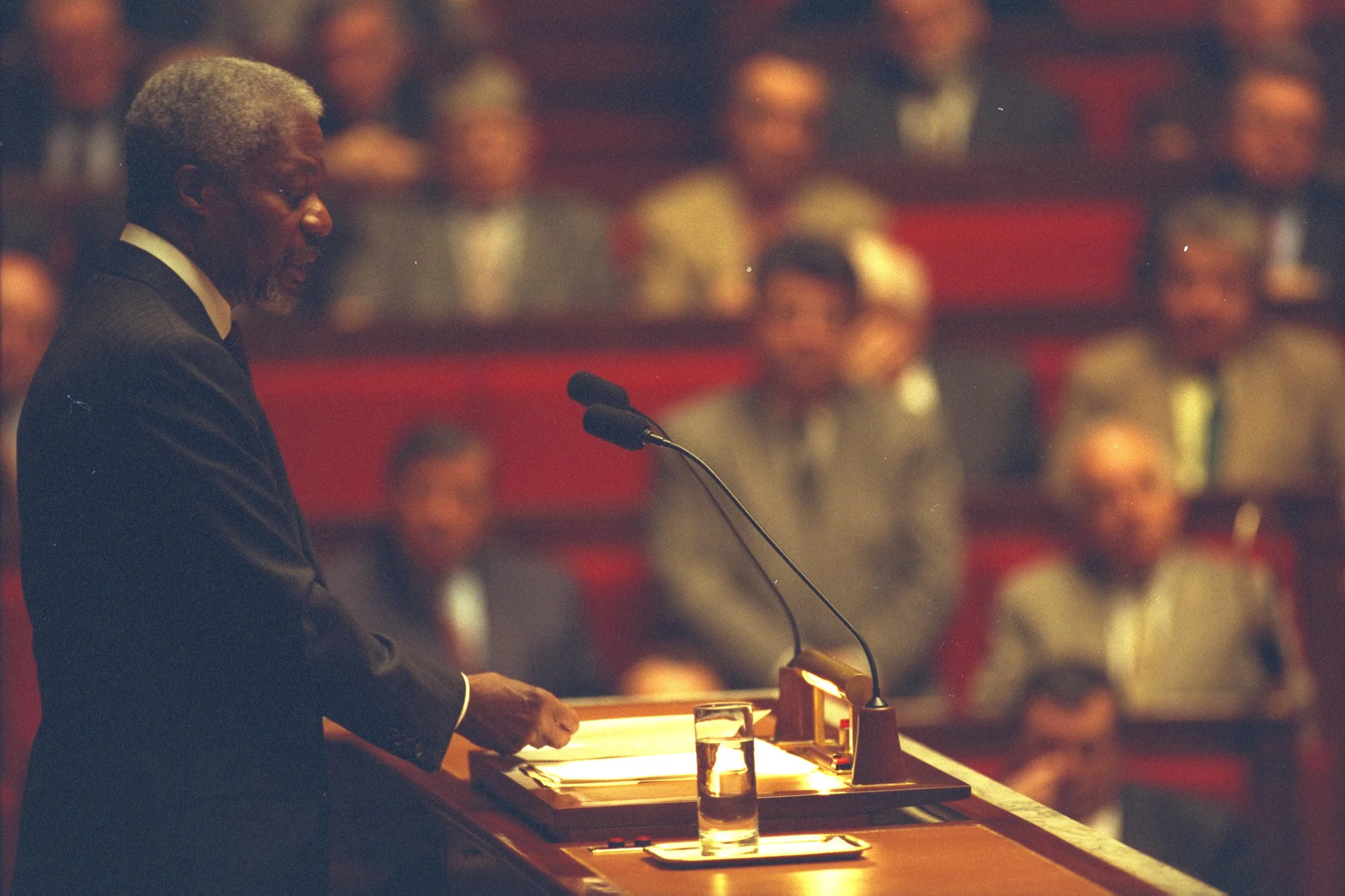 Kofi Annan speaks at a lectern with two microphones before a seated audience. The journalistic, action-oriented photo captures him in profile, emphasizing his role as a global leader in a formal, high-stakes setting.