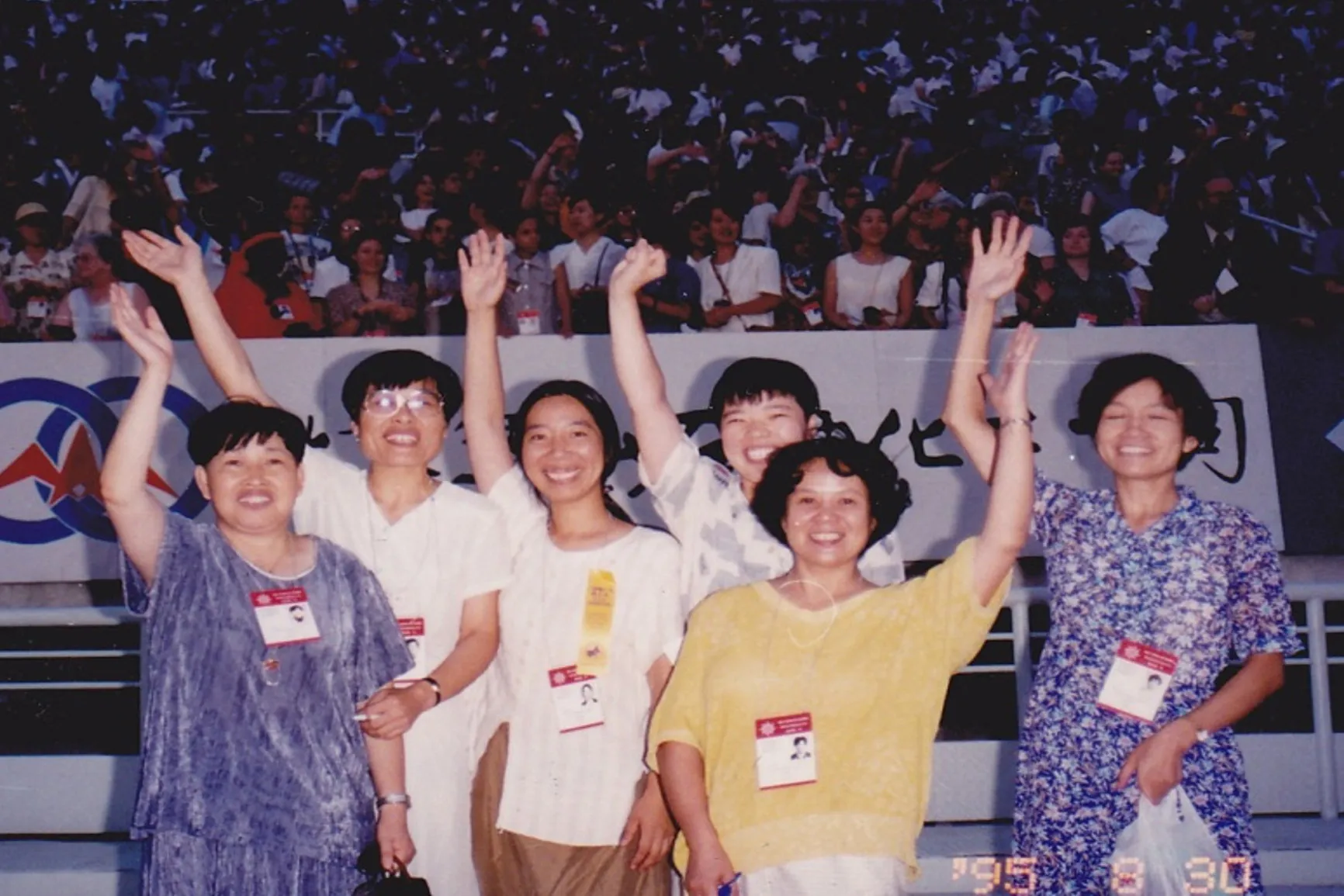 Six women stand with arms raised and joyful expressions in front of a stadium crowd and banner. This action-oriented, journalistic photo captures a moment of shared victory and humanizes the work of global advocates for social justice.