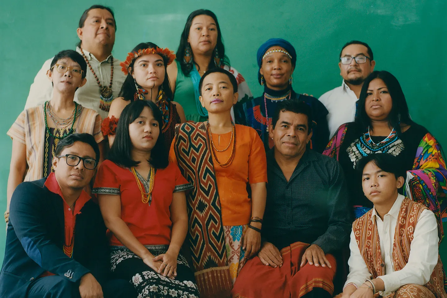 A portrait of a diverse group of 11 Indigenous leaders, including women and men in traditional patterned textiles and headpieces, posing together against a solid green background. The high-quality photograph humanizes the work of Indigenous communities.