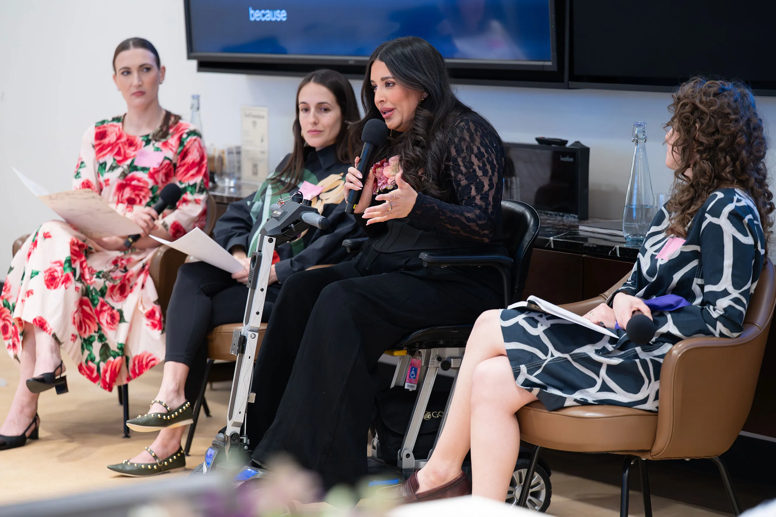 A group of four women participate in a panel discussion. One woman in a black lace top speaks into a microphone while using a mobility scooter, seated alongside other professionals. This image illustrates inclusive leadership, gender justice advocacy, and diverse perspectives at a high-level conference.