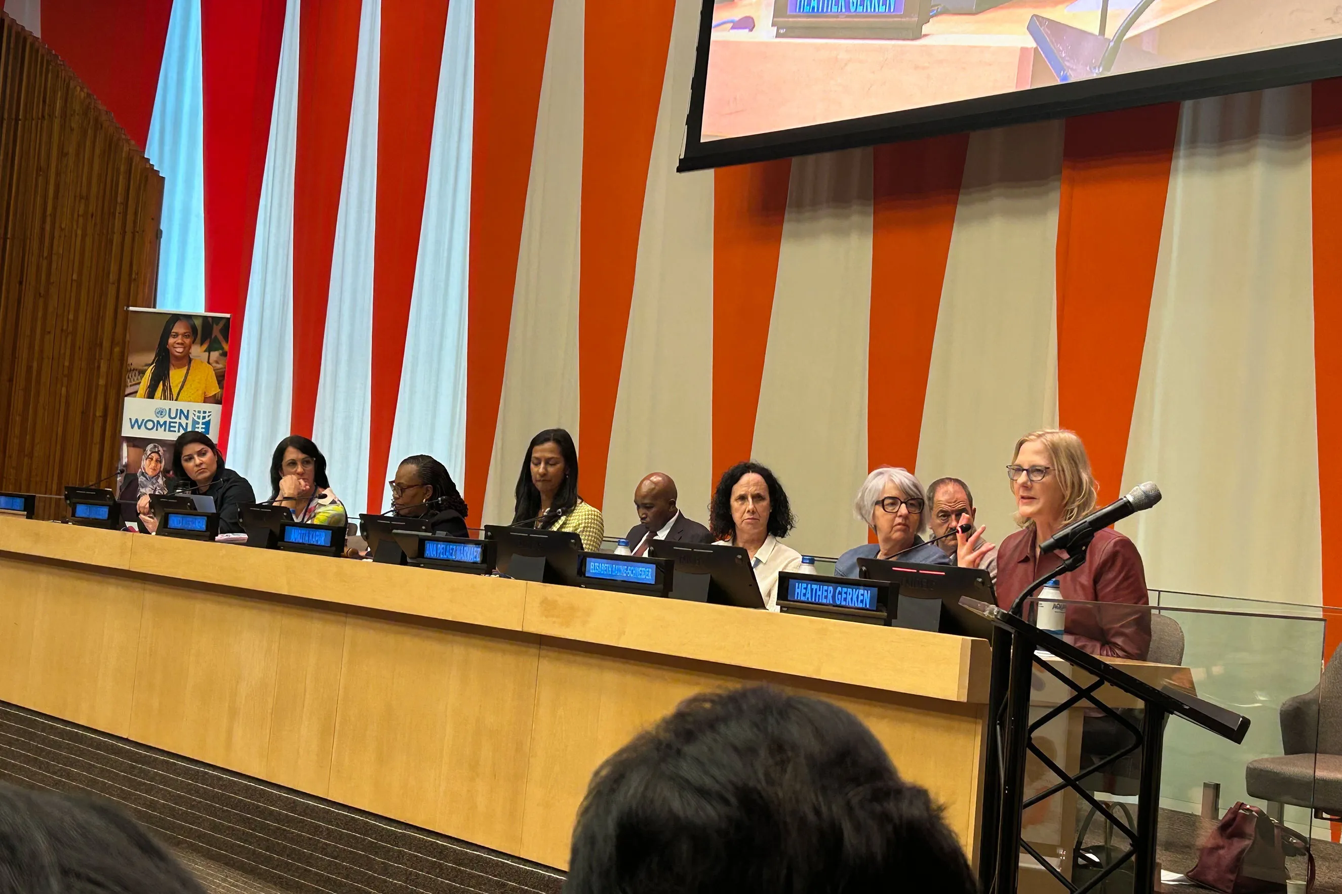 Heather Gerken sits at a long panel desk at a United Nations conference, speaking into a microphone. Behind her is a large UN Women sign, and other panelists are seated to her left. The image highlights international gender justice advocacy, policy discussion, and leadership.