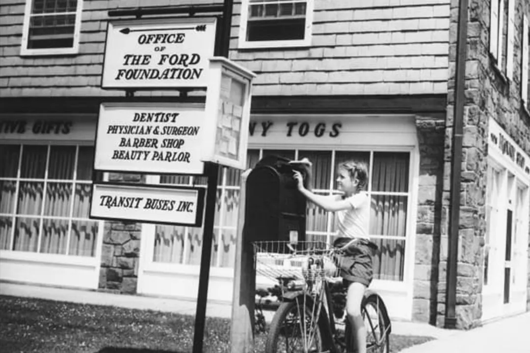 A vintage, black and white photograph from Middletown (Muncie, Indiana), depicting an inclusive community. A young girl on a bicycle uses a roadside mailbox. A sign in the background lists the local Ford Foundation office alongside essential medical services, illustrating civic participation and access for all residents.