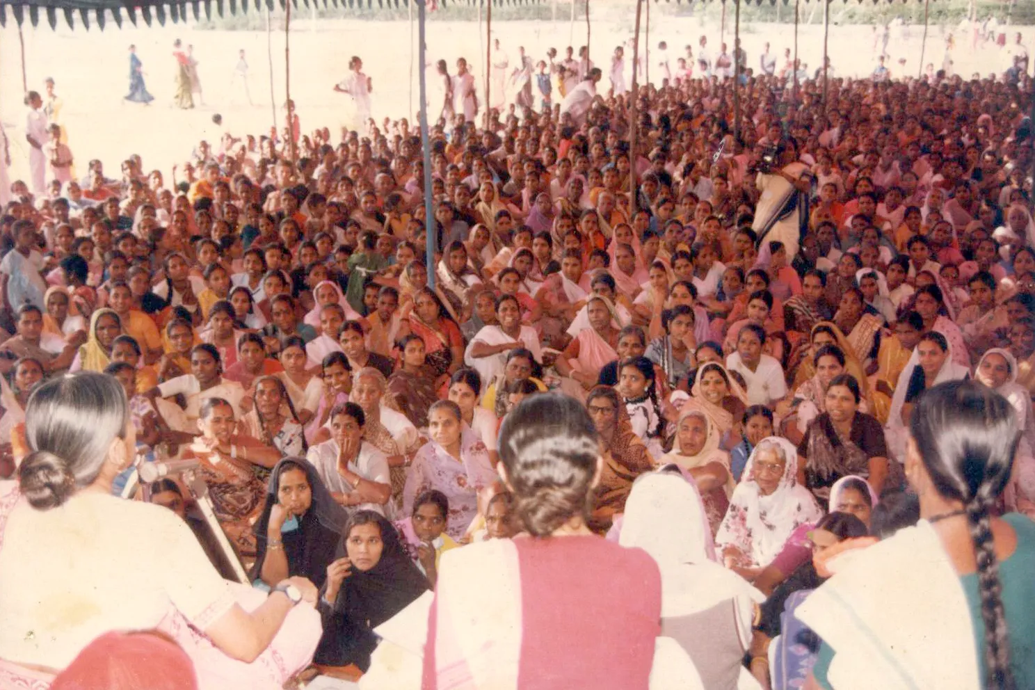 A high-angle shot shows a large, dense crowd of women seated on the ground under a tent in India. Most women wear colorful saris and face toward a stage in the foreground, where speakers are seen from behind addressing the gathering.