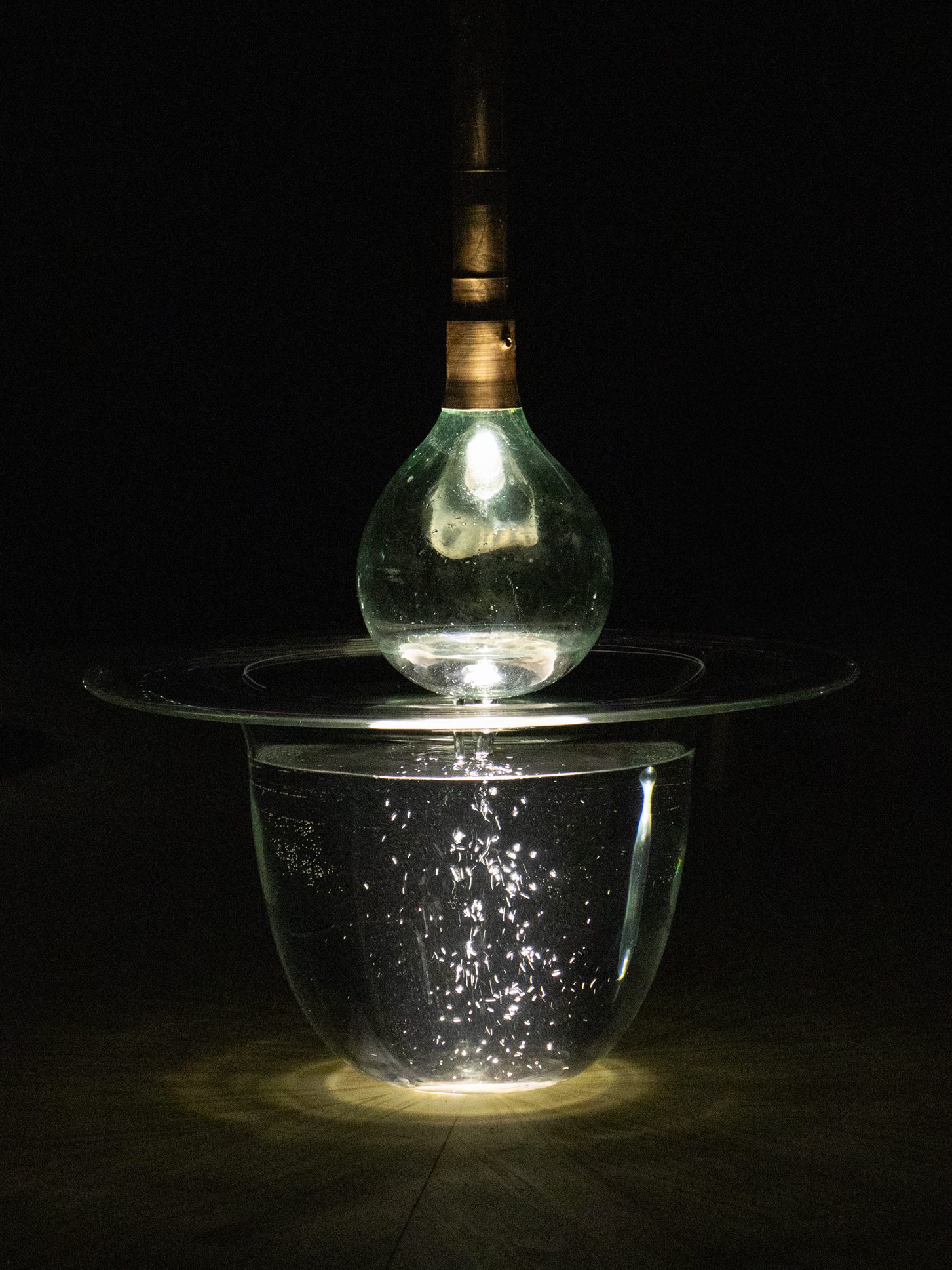 An illuminated light bulb attached to a copper rod is suspended above a glass bowl, both full of water.
