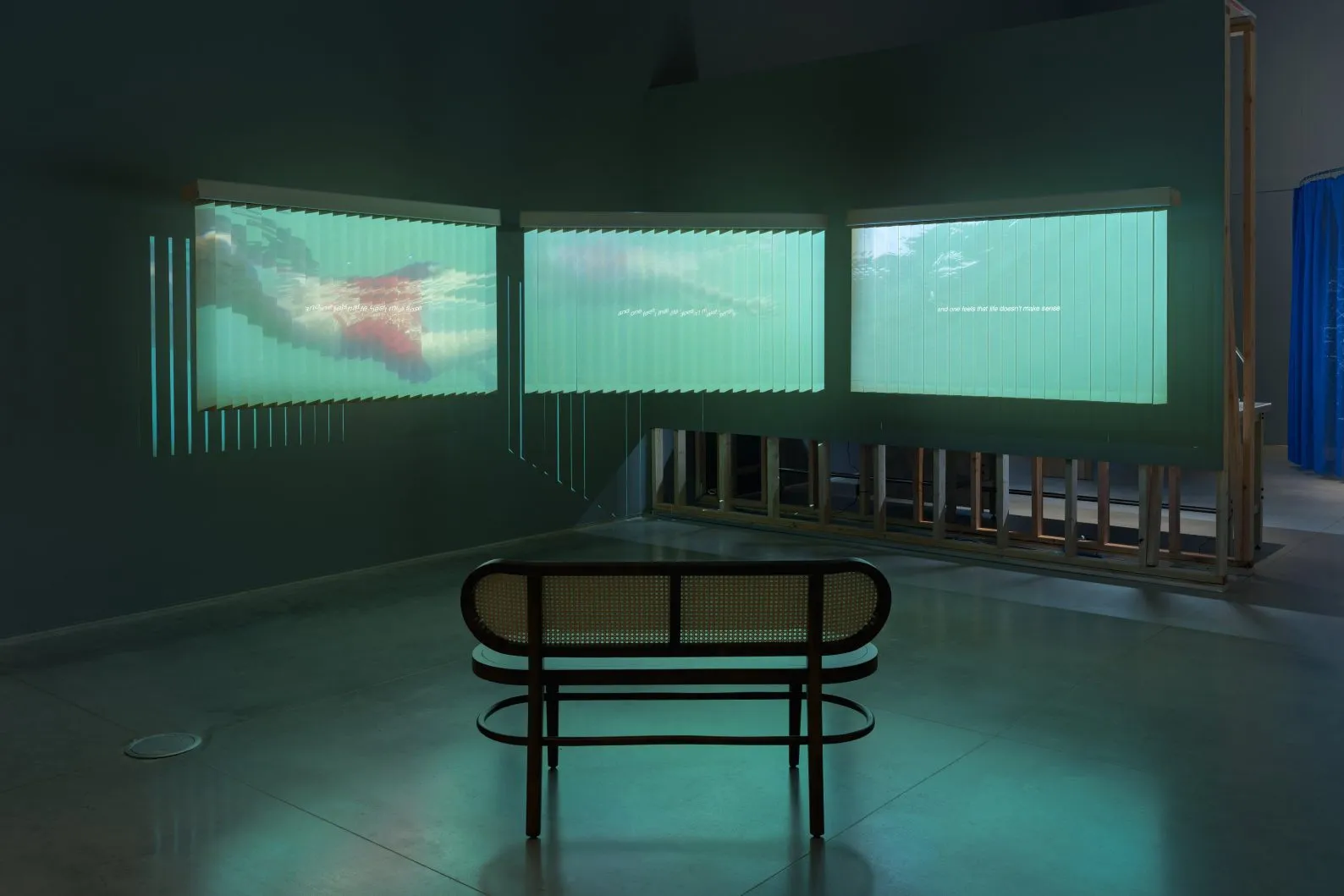 An installation featuring a three channel video projected onto vertical blinds as screens with a rattan bench in the center.