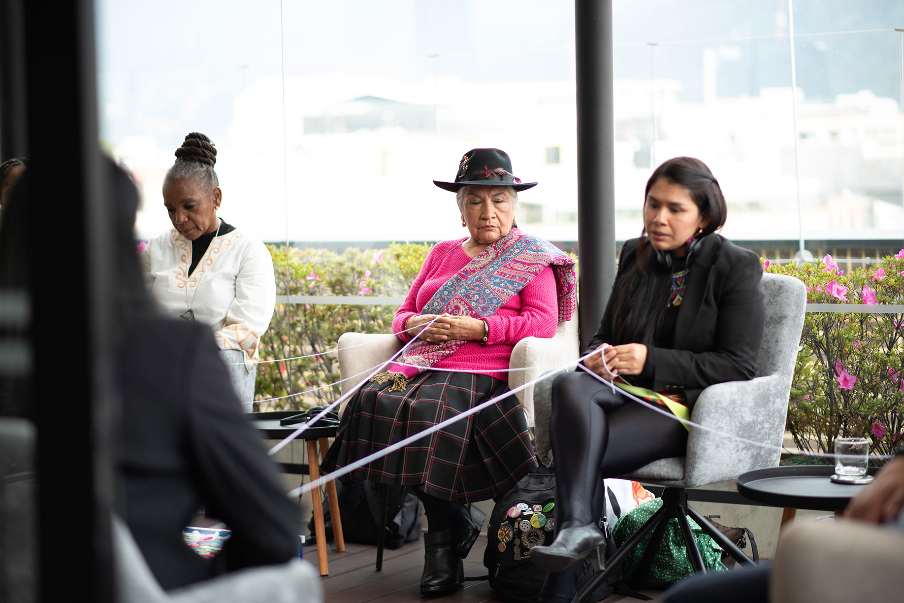 A group of people sitting in chairs indoors, engaging in a thread art activity. The person in the center wears a traditional hat and colorful attire. They are working with threads connected among them. The setting includes glass walls and potted plants.