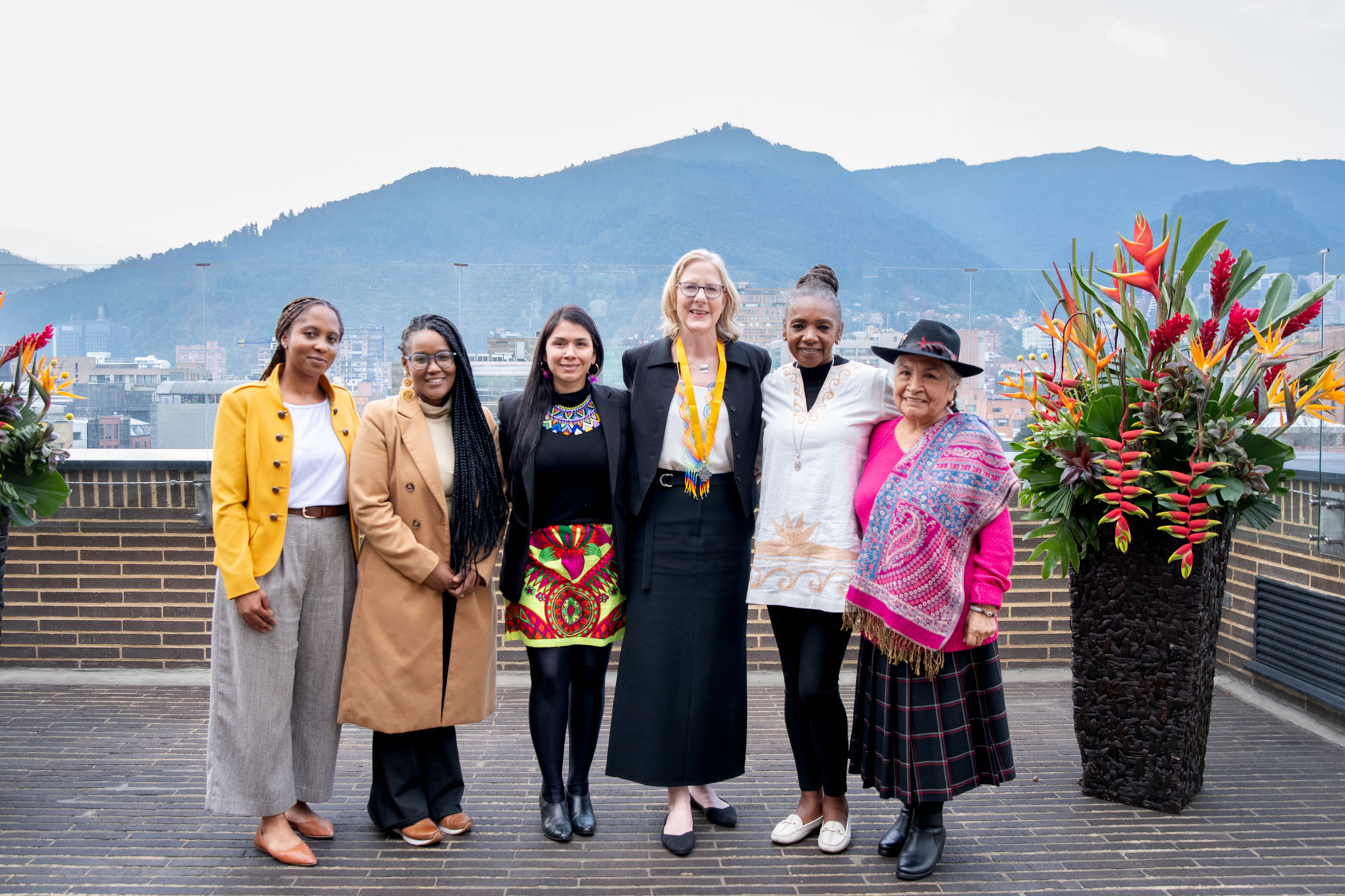 Heather Gerken, at the center of a group of people stands on a rooftop terrace with two large floral arrangements on either side. In the background are mountain peaks and a cityscape.