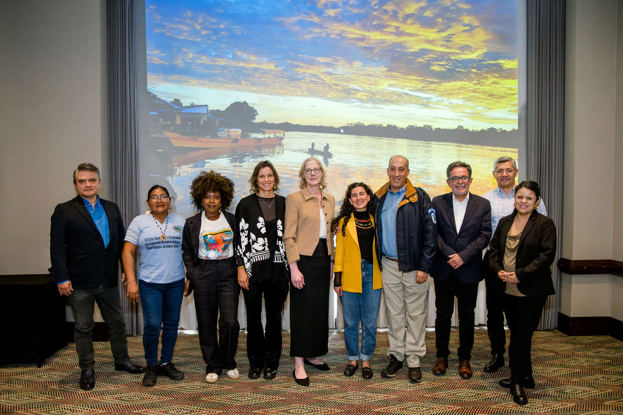 Heather Gerken at the center of a group of people standing in front of a projection screen displaying a sunset river scene. They are posed in a row, smiling at the camera. The setting is a meeting room.