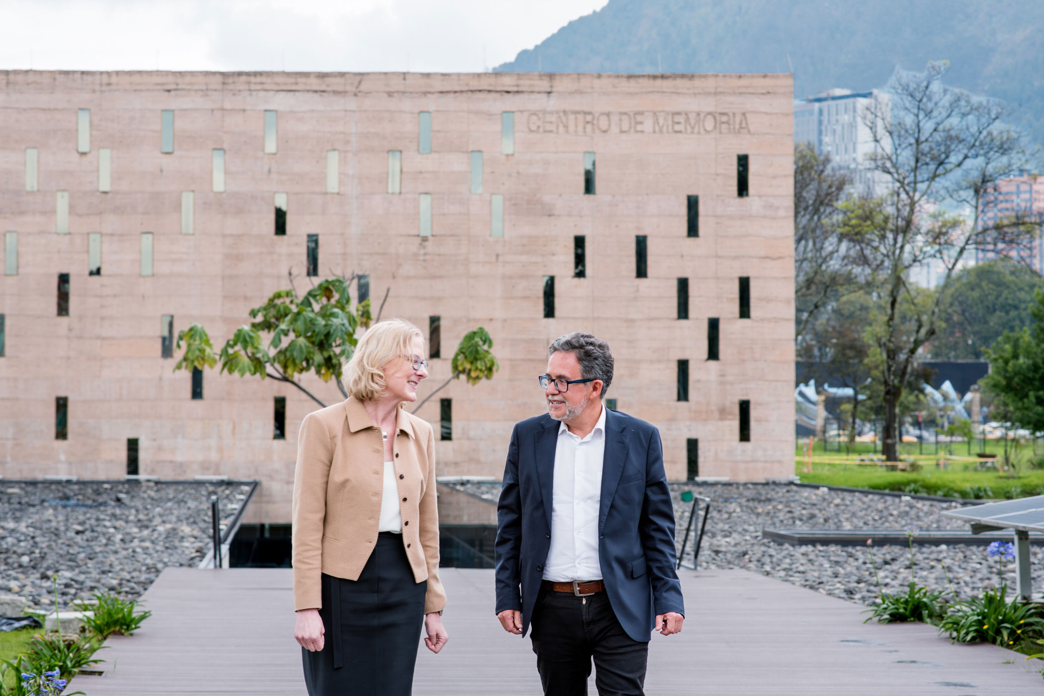 Heather Gerken and  Javier Ciurlizza walk along a path in front of the Centro de Memoria, Paz y Reconciliación building. They appear engaged in conversation, exuding a professional and friendly tone.