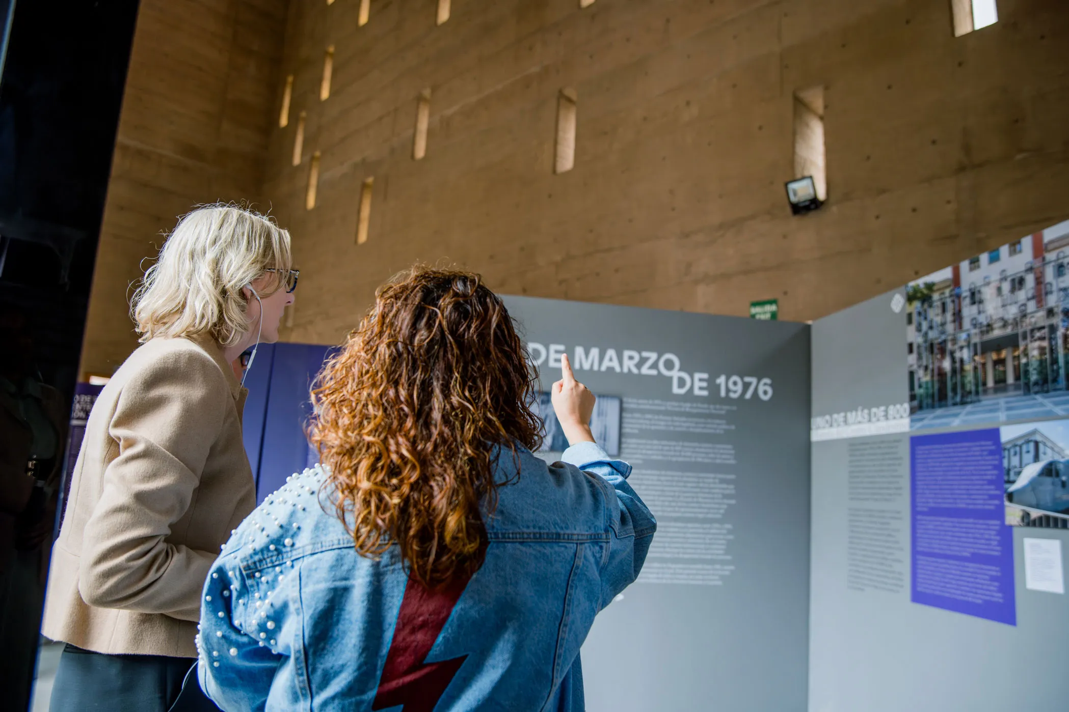 Heather Gerken and an individual are observing an exhibition display with text about events from March 1976 at the Center of Memory, Peace, and Reconciliation. The setting appears to be an indoor gallery with high ceilings and narrow windows. One person gestures towards the exhibit. The focus is educational and historical, serving as a space to honor the millions of victims of the country's internal armed conflict.
