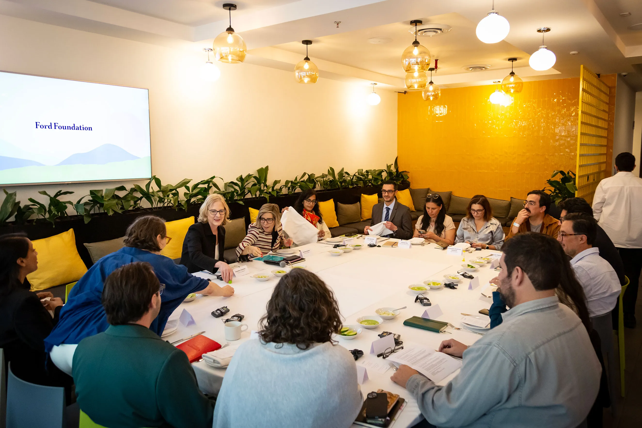 A photo of professional staff members gathered around a large white table in a vibrant meeting room with yellow accents. A screen displays the Ford Foundation logo. This journalistic image captures a moment of collaborative dialogue and humanizes the foundation's regional work.