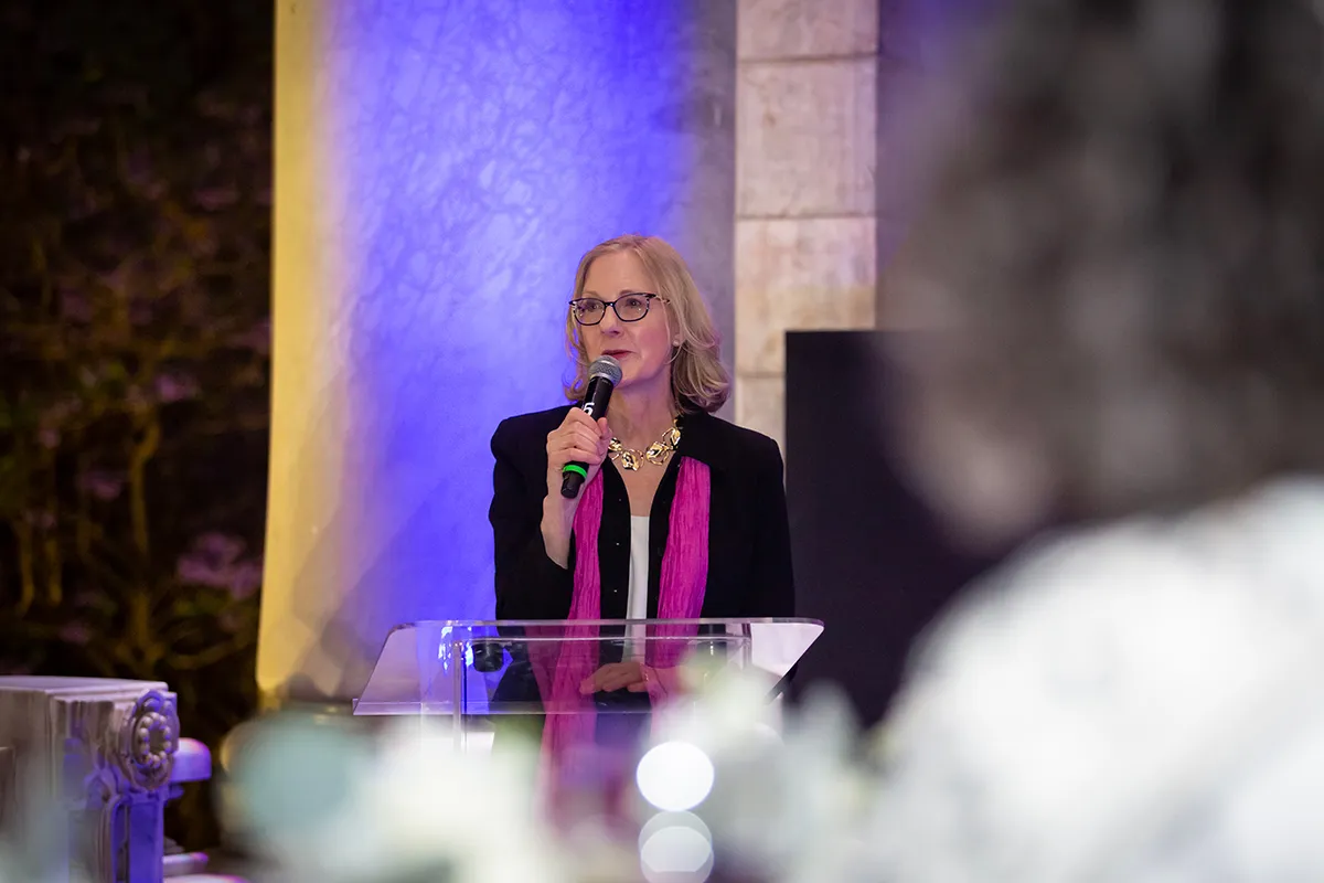 Heather Gerken in glasses and a pink scarf speaks at a transparent lectern during an evening event. This high-quality, action-oriented photo captures a moment of leadership and communication, humanizing the foundation’s work in a professional regional setting.