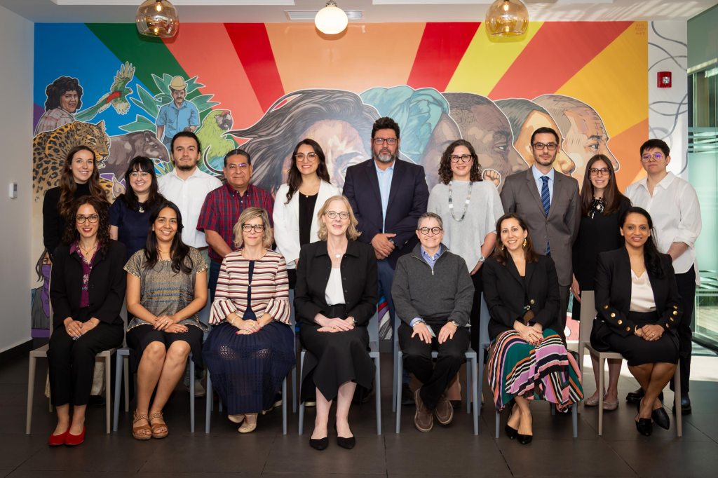 A group portrait of 16 professional staff members posing in two rows in front of a colorful, vibrant mural in Mexico City. The diverse team is seated and standing together, humanizing Ford’s collaborative work with regional partners.